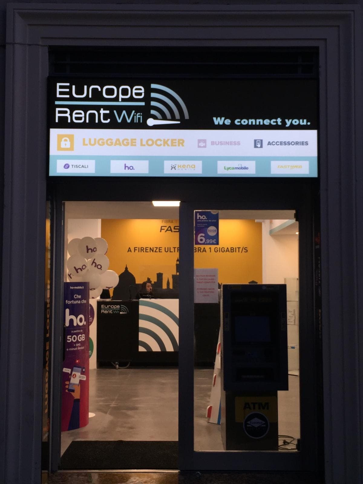 L’insegna e l’ingresso di Europe Rent WiFi a Largo Fratelli Alinari 27, Firenze, illuminati di sera